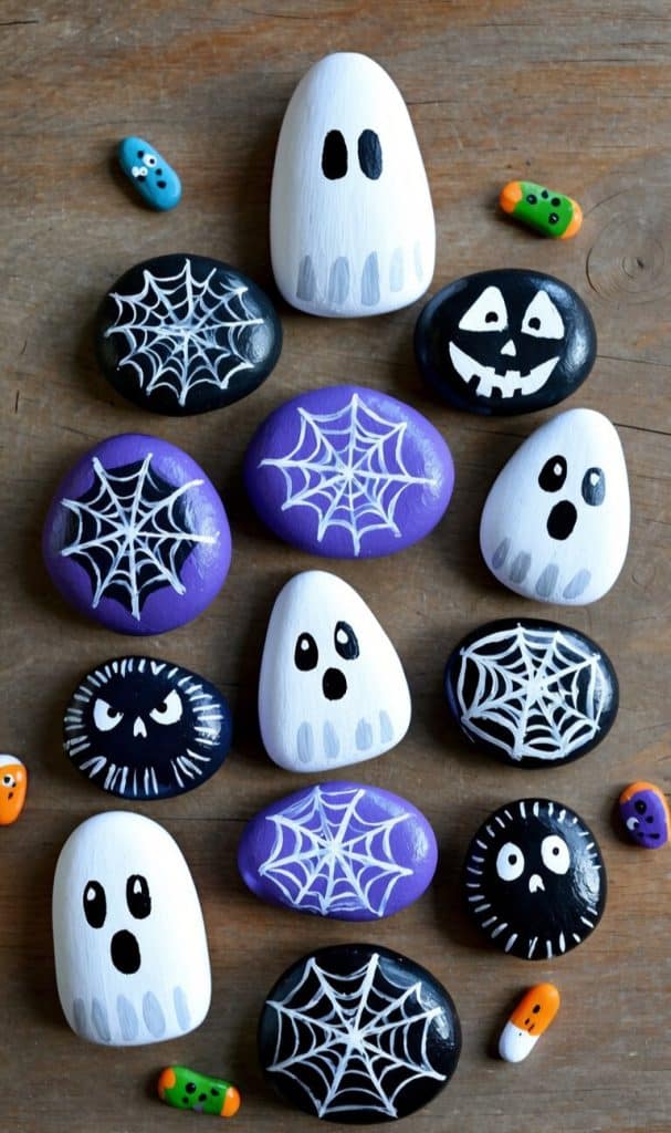 Painted rocks with Halloween designs, featuring ghost painted rock ideas, black and purple rocks with spiderwebs and spooky faces, are scattered on a wooden surface. Small, colorful candy-shaped decorations surround the rocks.