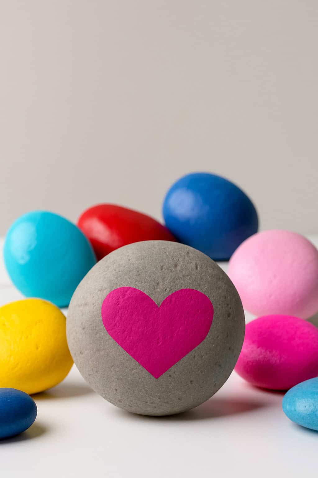 A gray stone with a large pink heart painted on it is surrounded by colorful stones in blue, red, yellow, and pink on a white surface with a light background.