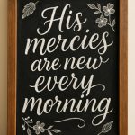 A framed blackboard with white chalk lettering reads, "His mercies are new every morning," decorated with hand-drawn flowers and leaves in the corners. The frame rests on a wooden surface.