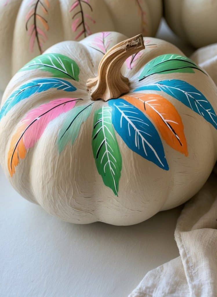 A white pumpkin decorated with colorful painted leaves in shades of green, blue, pink, and orange surrounding the stem. The pumpkin rests on a light surface next to a beige cloth.