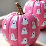 Two pink pumpkins decorated with cute white ghost illustrations are sitting on a light surface in natural sunlight. The background is softly blurred, emphasizing the playful, Halloween-themed pumpkins.