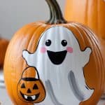 A pumpkin painted with a smiling white ghost holding a jack-o'-lantern bucket. The ghost has rosy cheeks and black eyes, with another painted pumpkin visible in the background. Candy corn pieces are on the table.