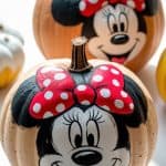 Two pumpkins are painted with large Minnie Mouse faces, each featuring a red bow with white polka dots, big eyes, and a smiling expression. Sunlight highlights the pumpkins on a white surface.