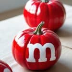 Three pumpkins painted shiny red with a large white "m" on each, resembling red M&M candies, are displayed on a light surface.