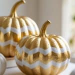 Two decorative pumpkins painted gold with white zigzag stripes sit on a white surface, with a soft, blurred background.