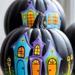 Two black pumpkins are painted with bright, whimsical houses in blue, purple, and yellow, featuring arched doors and windows. The pumpkins are stacked together on a light surface.