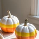 Two pumpkins painted in white, yellow, and orange stripes resembling candy corn sit on a white surface near a sunlit window.
