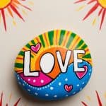 A painted rock with the word "LOVE," colorful patterns, and hearts is placed on a surface decorated with bright, stylized sun illustrations.