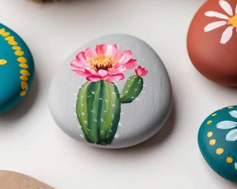 A smooth gray stone painted with a green cactus and a pink flower, surrounded by other colorful painted stones on a white surface.