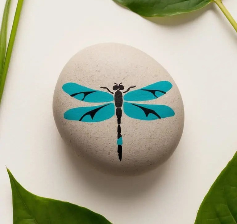 A smooth, round stone painted with a blue and black dragonfly design, placed on a white surface surrounded by green leaves.